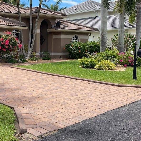 A brick driveway leads to a beige house with a brown roof, surrounded by green grass and landscaping.