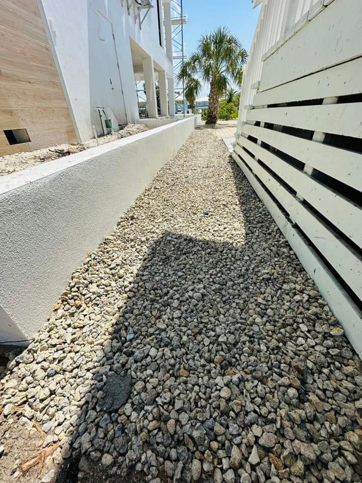Gravel pathway between white buildings, with sunlight casting shadows. Palm tree visible in the distance under a bright sky.