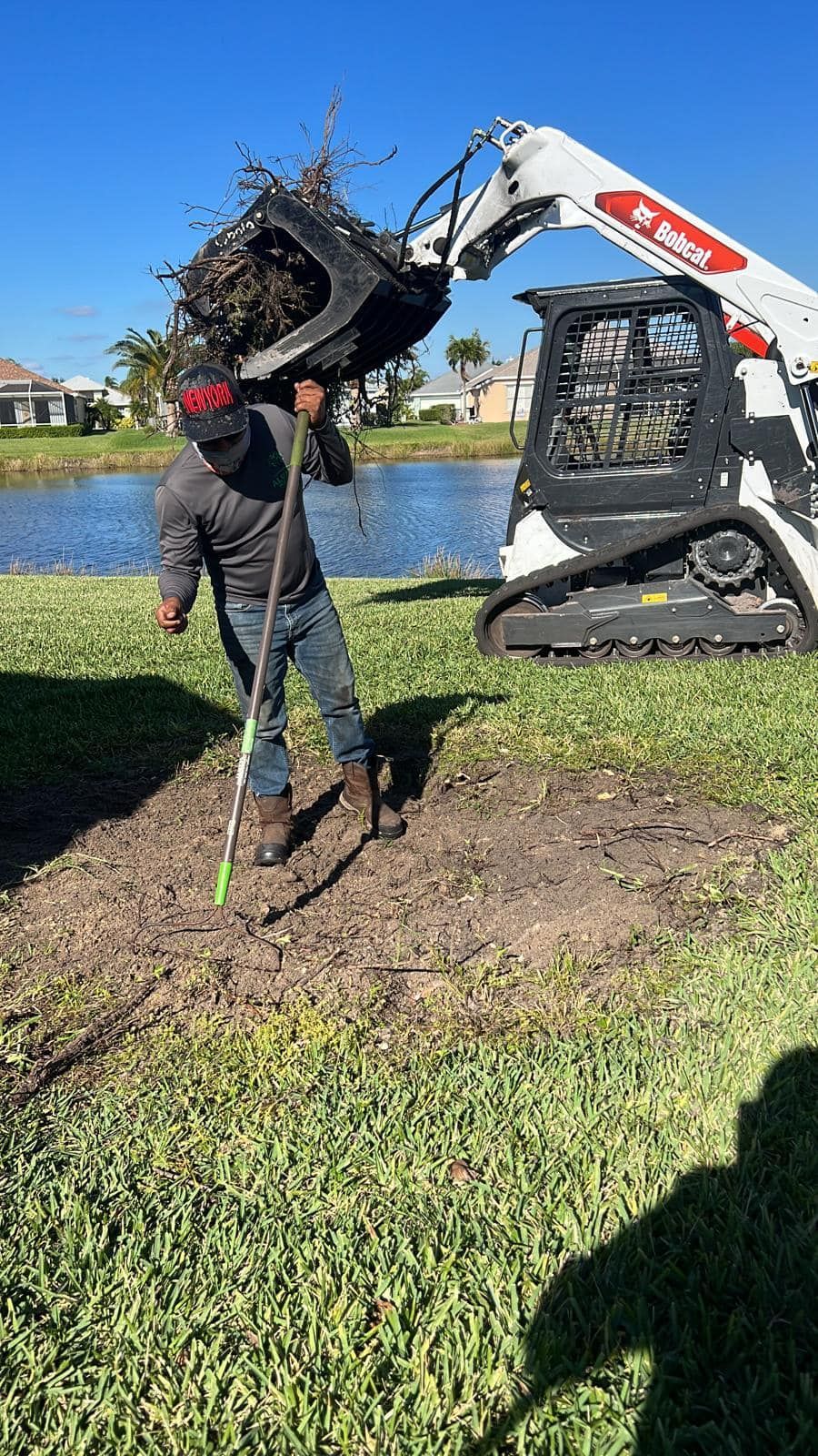 Man raking a dirt patch near a pond while a Bobcat holds debris; sunny day.