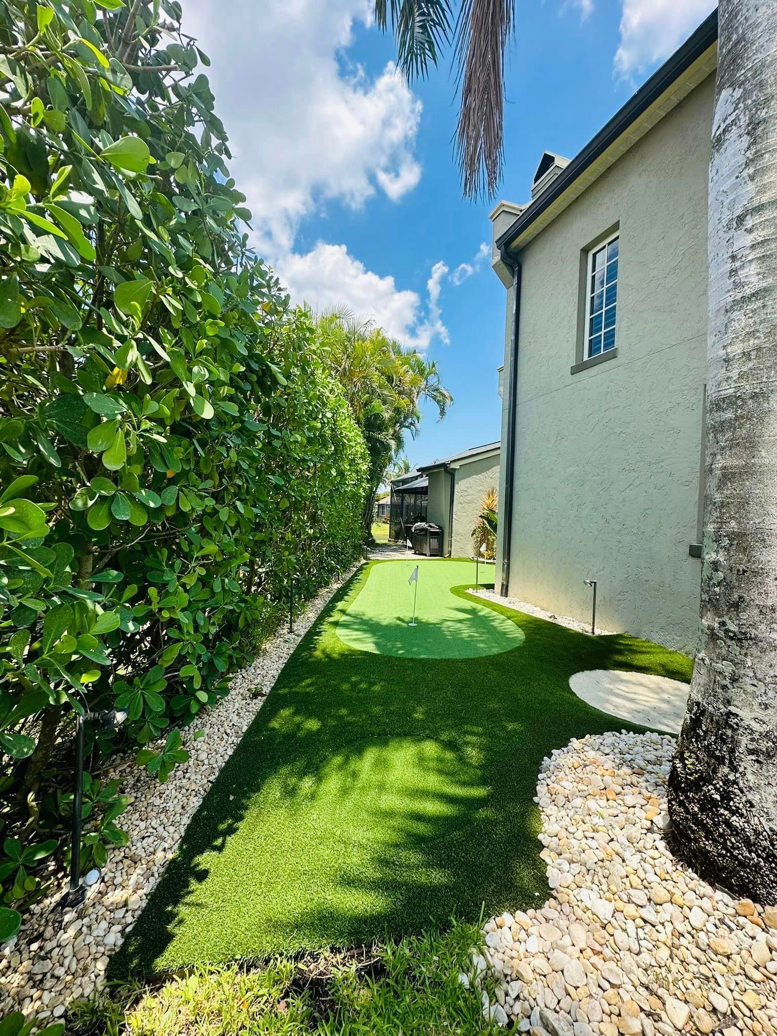 A narrow backyard with a putting green, hedges, and a light-colored building under a blue sky.