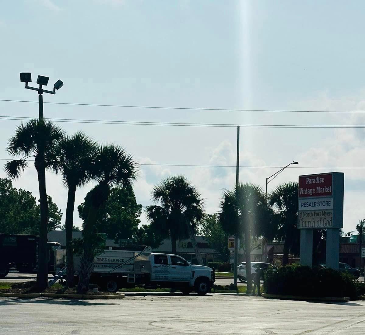 Exterior view of a parking lot on a partly cloudy day with a sign for various businesses, a pickup truck, and palm trees.