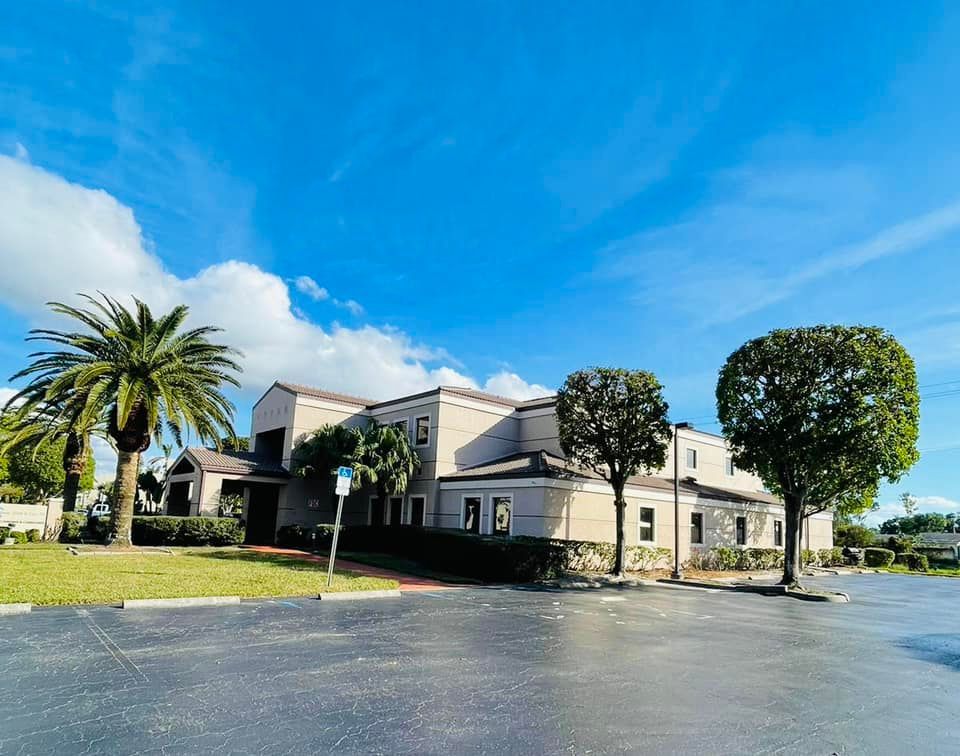 Exterior view of a two-story building with beige walls, a red brick walkway, and a blue sky with palm trees in front.