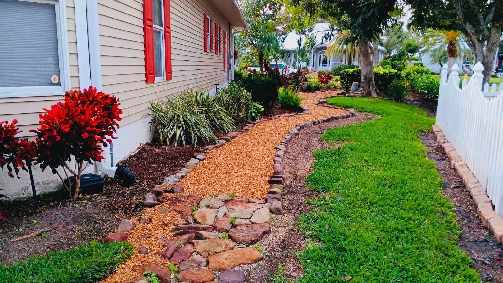 A winding gravel path lined with plants and a white picket fence beside a beige house with red shutters.
