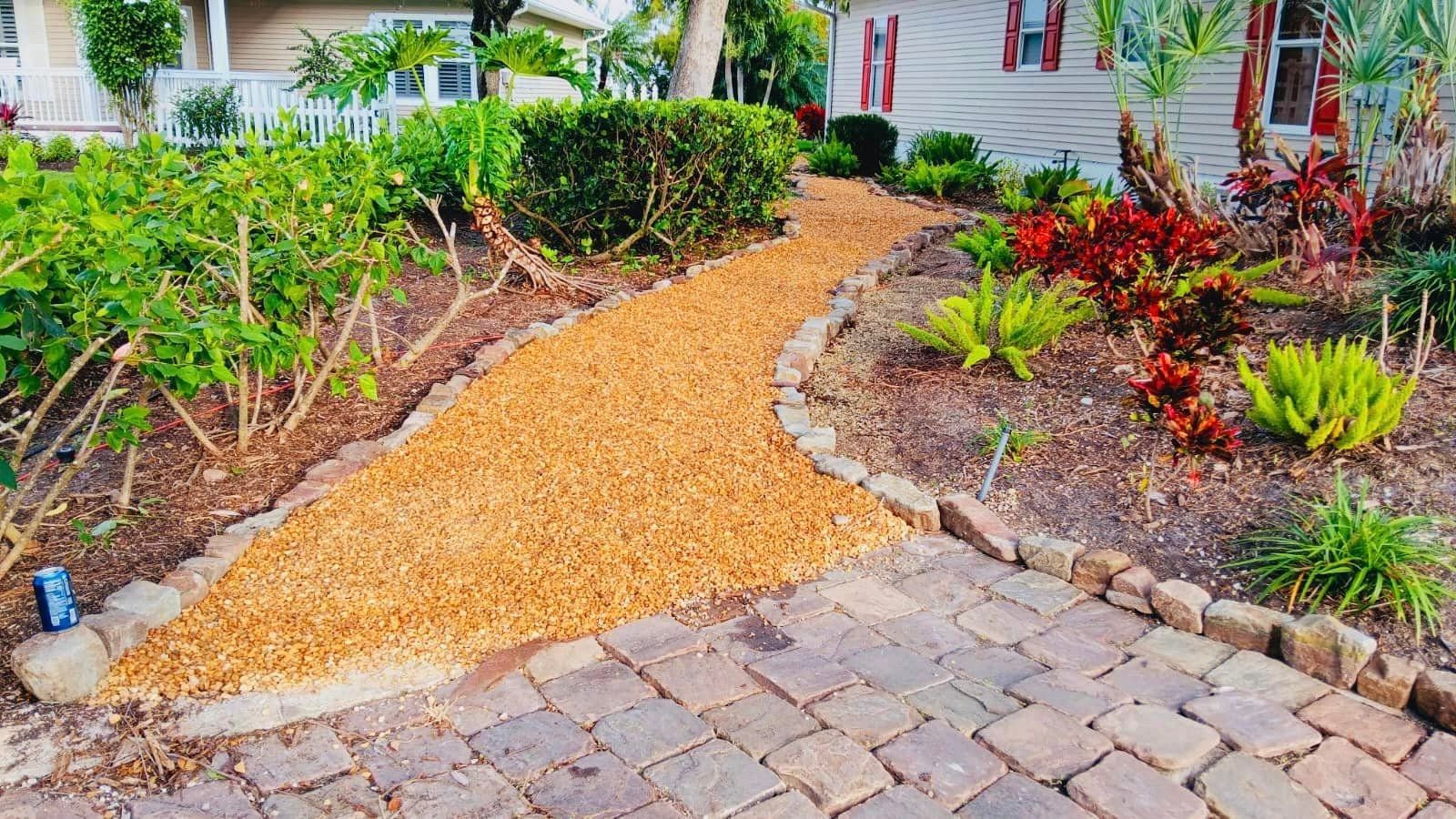 A gravel pathway curves through a well-landscaped yard, bordered by brick and colorful plants, leading towards a house.