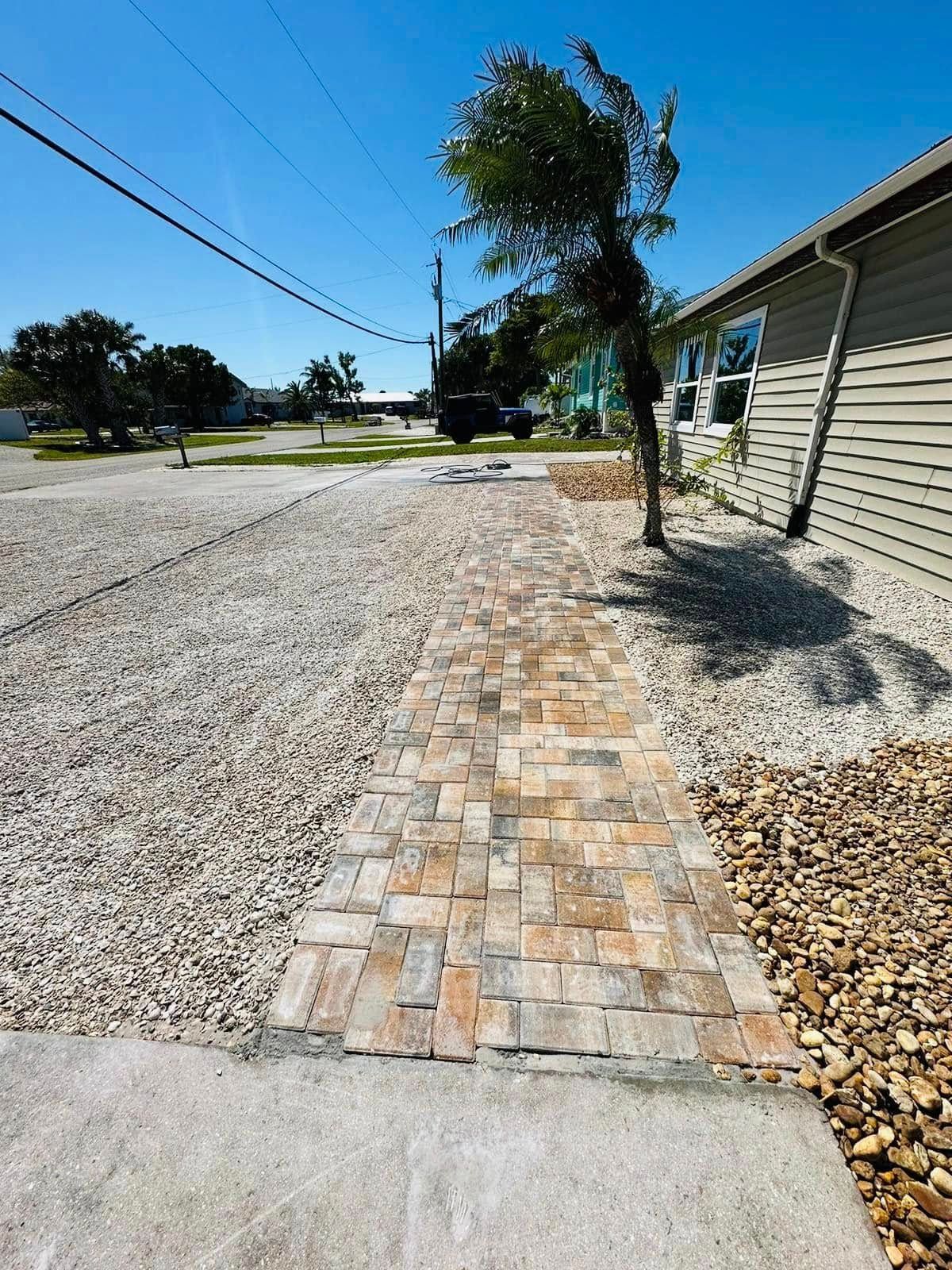 A brick pathway leads towards a house with tan siding, with gravel and small rocks on either side.