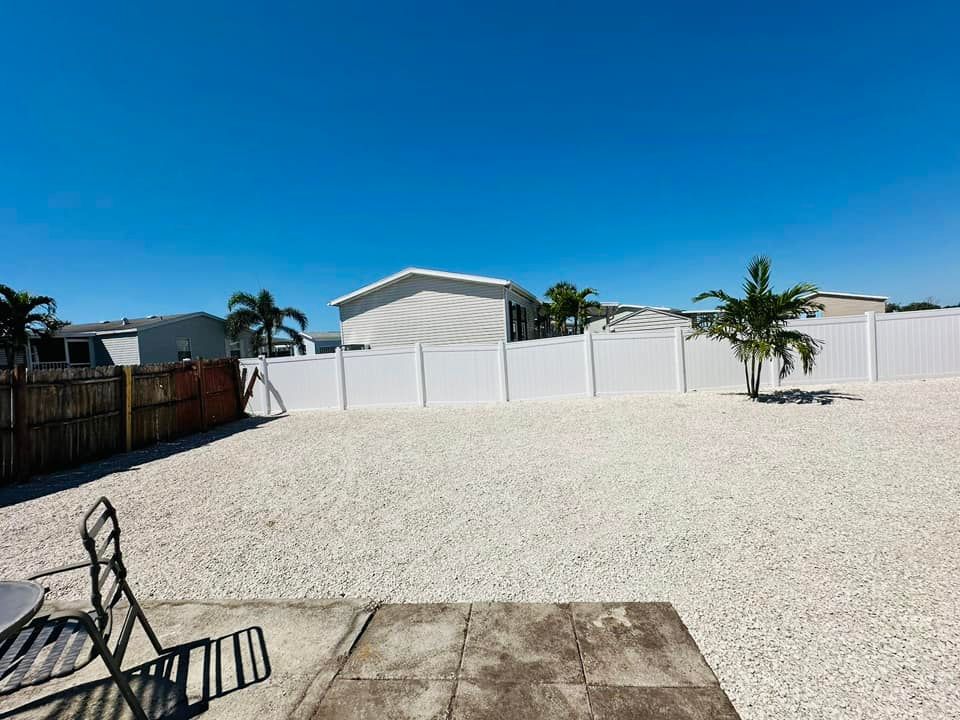 A backyard with white gravel, a white fence, and palm trees under a bright blue sky.
