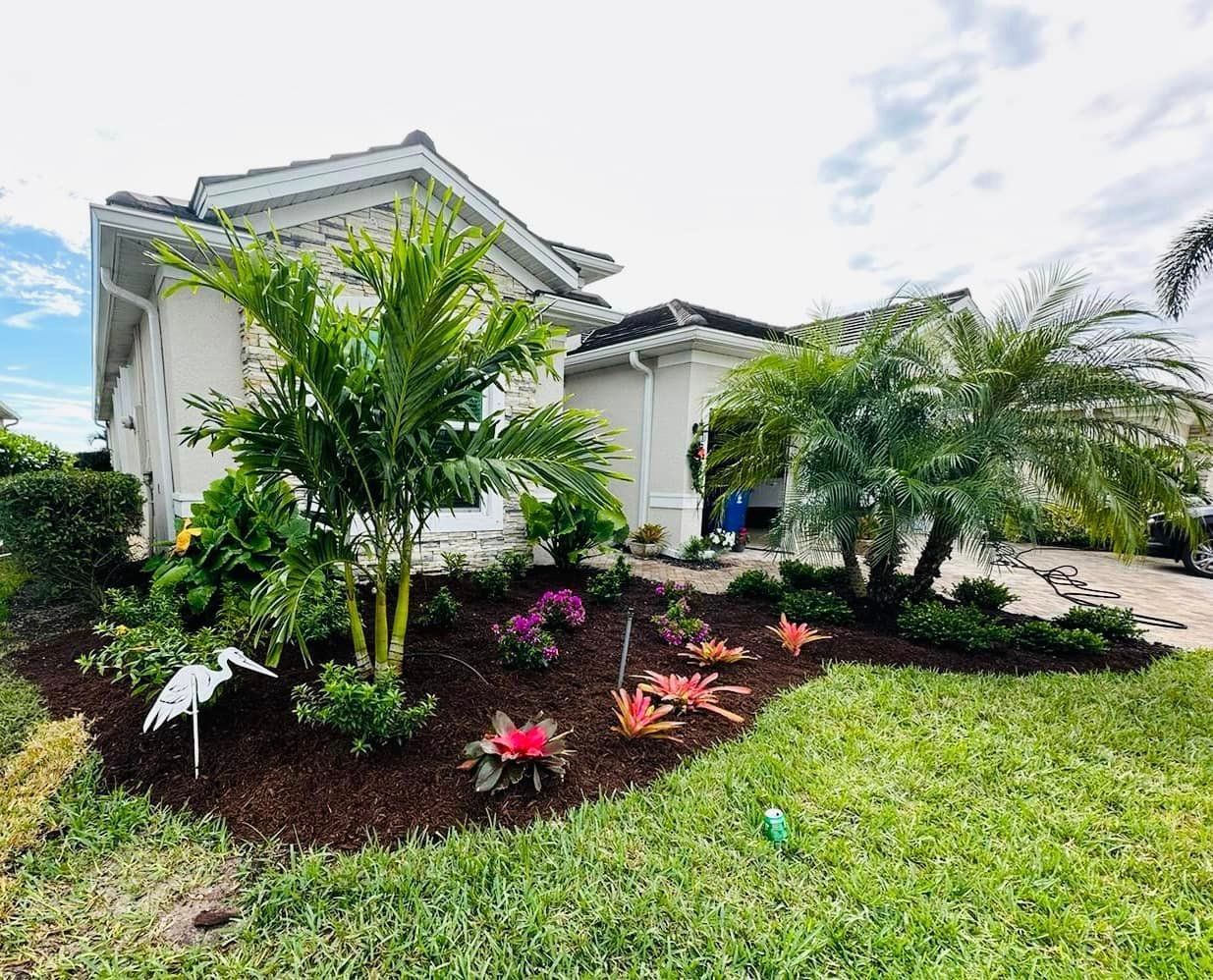 A beige house with lush tropical landscaping, including palm trees and colorful flowers, in front of a green lawn.