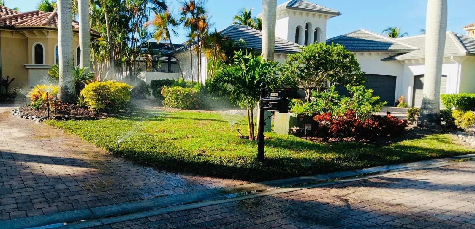 Lush green lawn and landscaping in front of a white house, watering by sprinklers on a sunny day.