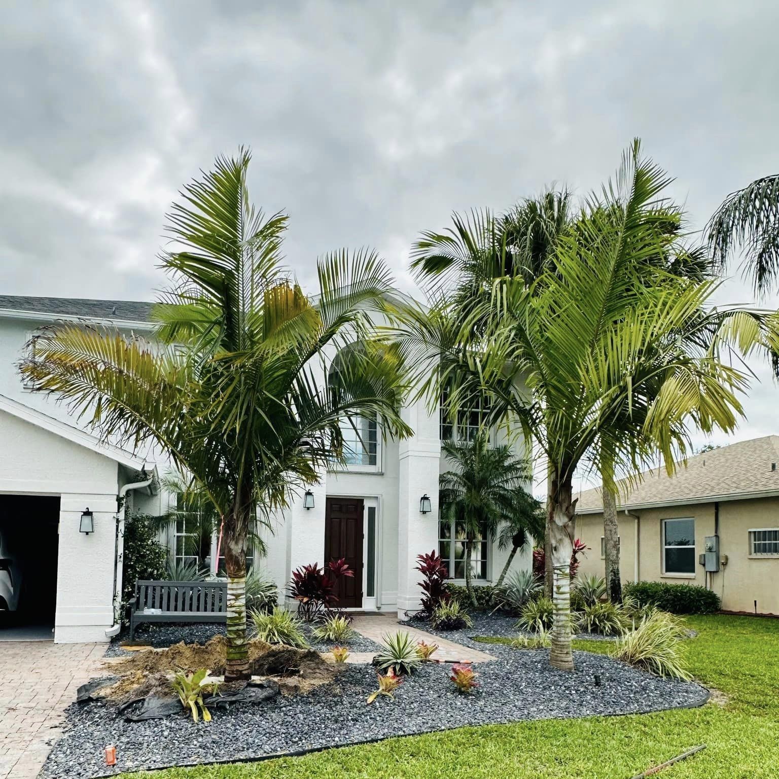 White two-story house with palm trees and a dark gray rock landscape, with a cloudy sky.