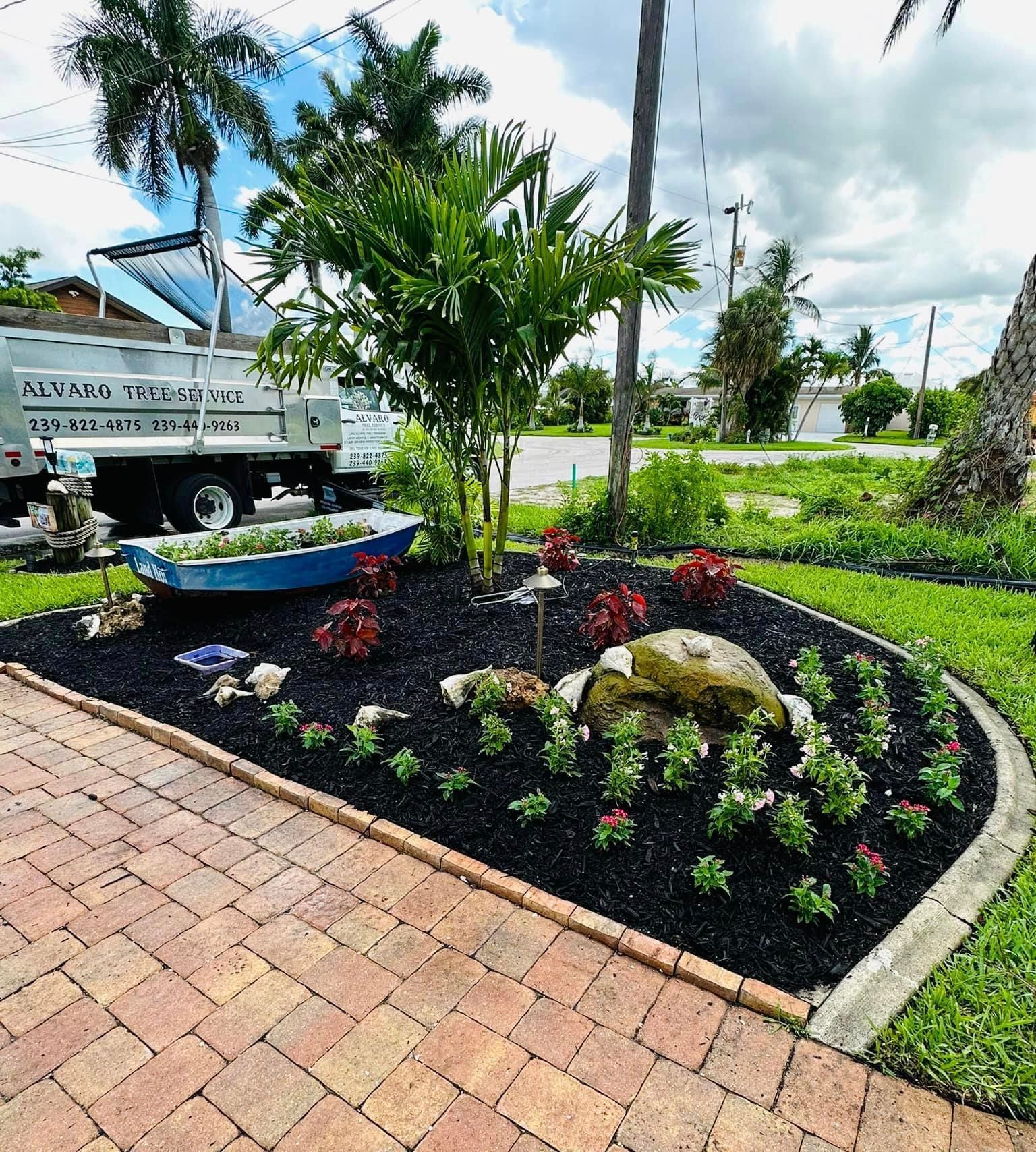 Landscaped garden bed with black mulch, colorful flowers, a rock feature, and a small blue boat, next to a brick pathway.