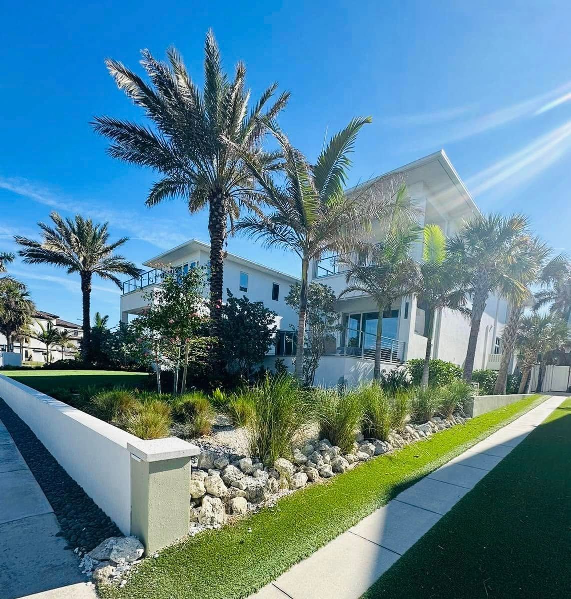 White beach house with palm trees on a sunny day. Green grass and a stone wall are in the foreground.