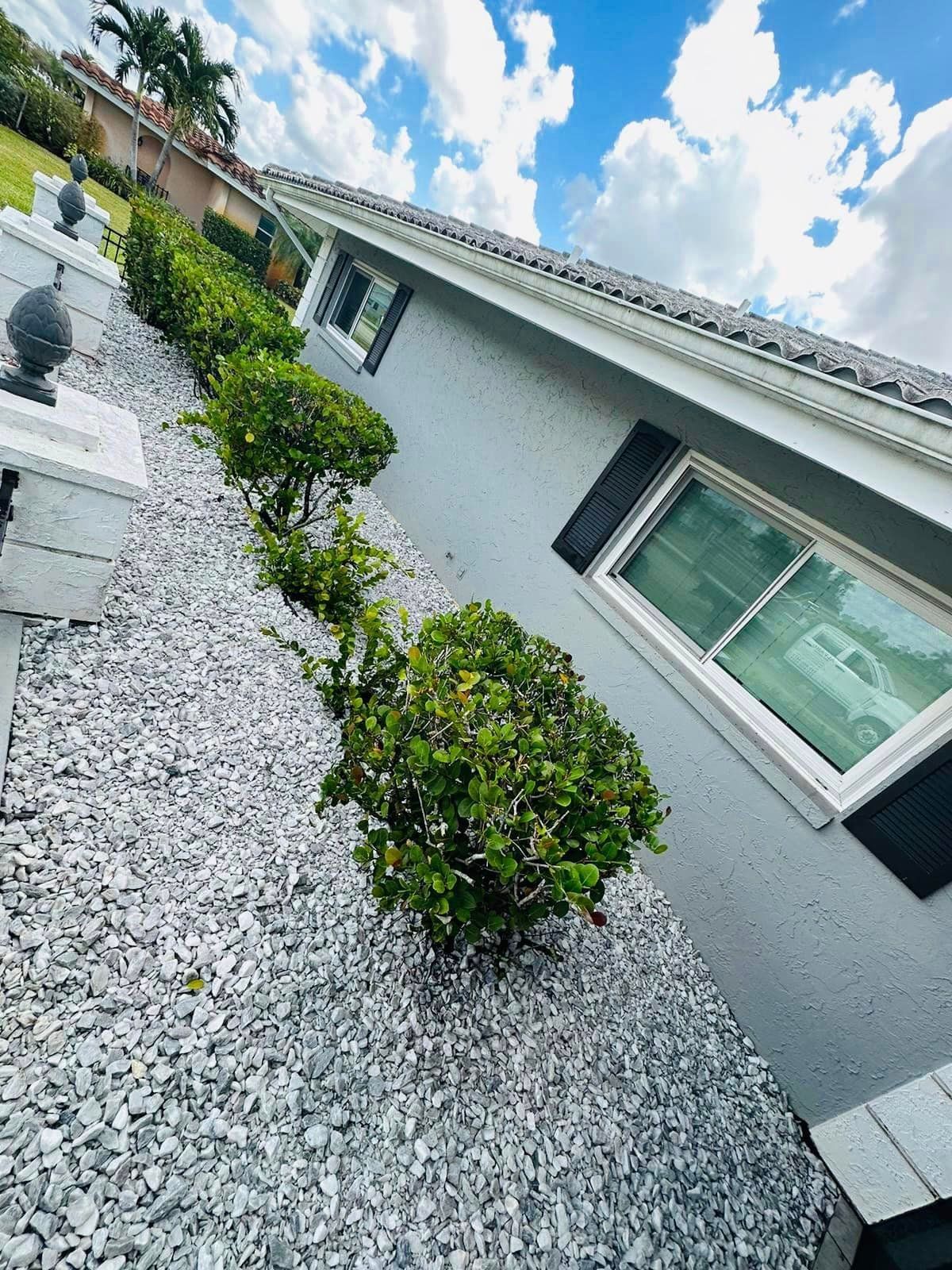 A house with a gray exterior and black shutters has a landscaped bed of white gravel and green bushes on a slope.