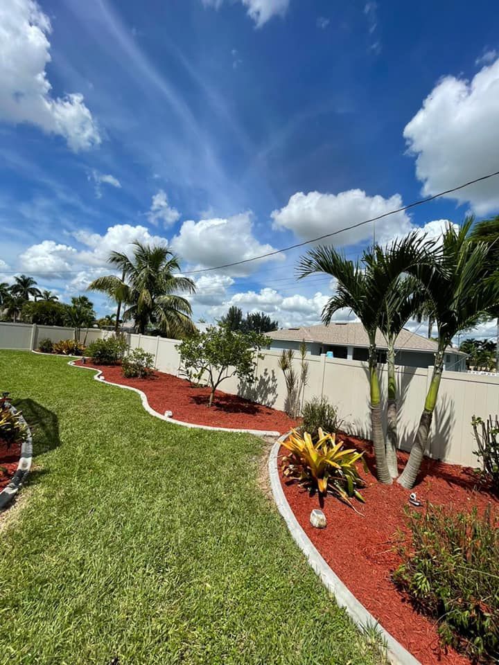 Lush green lawn and red mulch border a white wall with palm trees, under a partly cloudy blue sky.