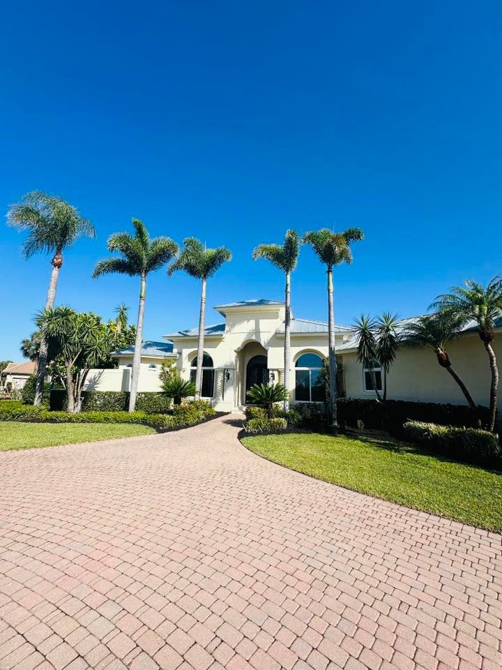 A large, light-colored house with a curved driveway lined with palm trees under a bright blue sky.