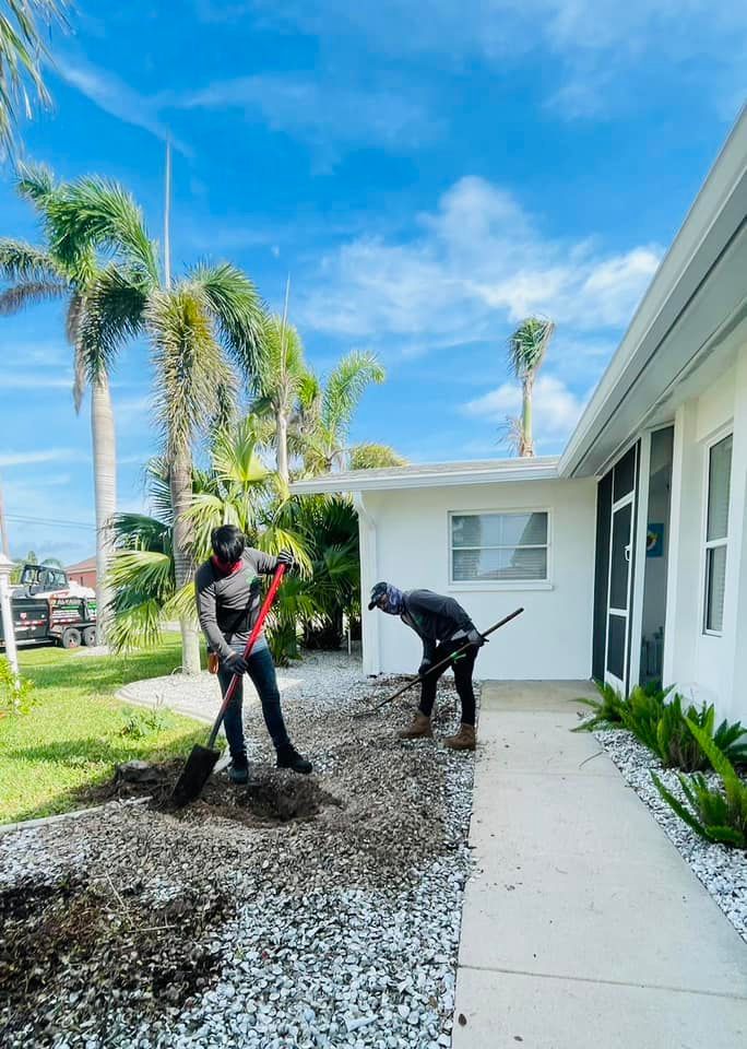 Two workers are shoveling dirt and gravel outside a white house on a sunny day with palm trees in the background.