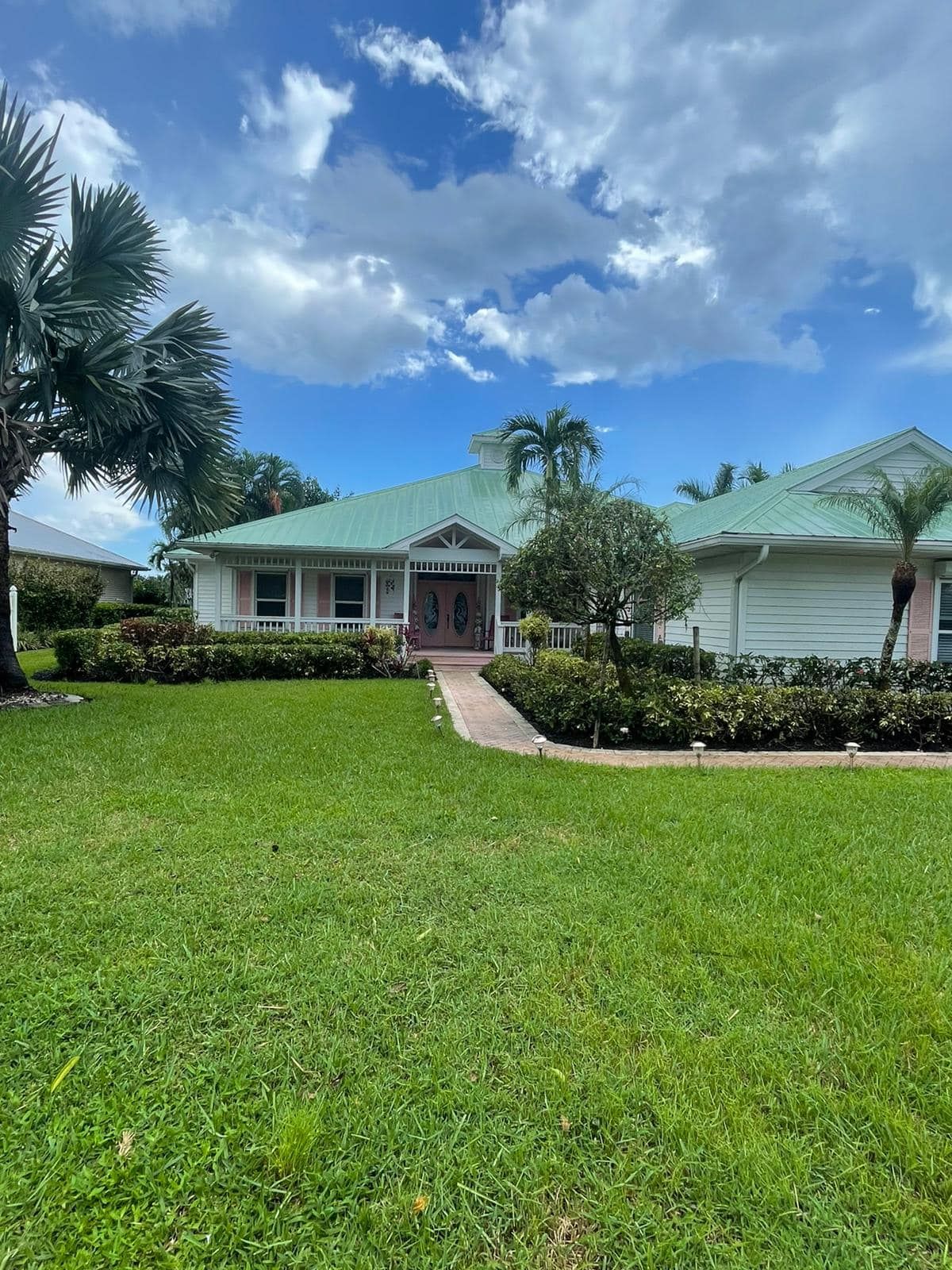 A white house with a green roof sits on a lawn. Palm trees frame the house under a partly cloudy sky.