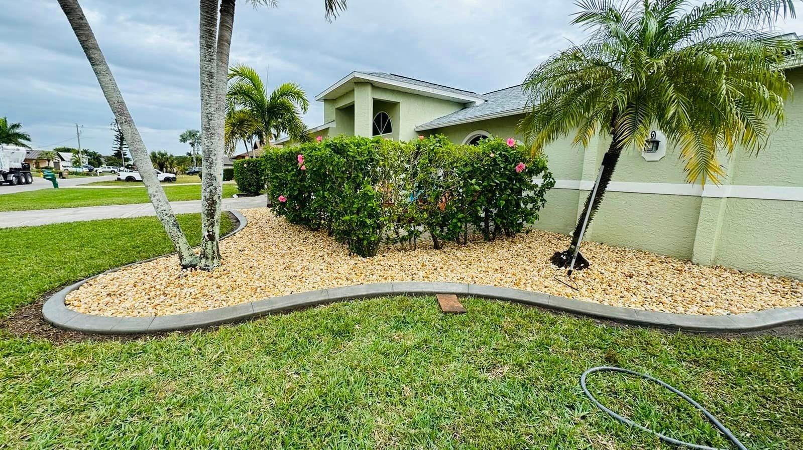 Landscaped front yard with tan rock bed, green shrubs, and a light green house with a curved concrete border.