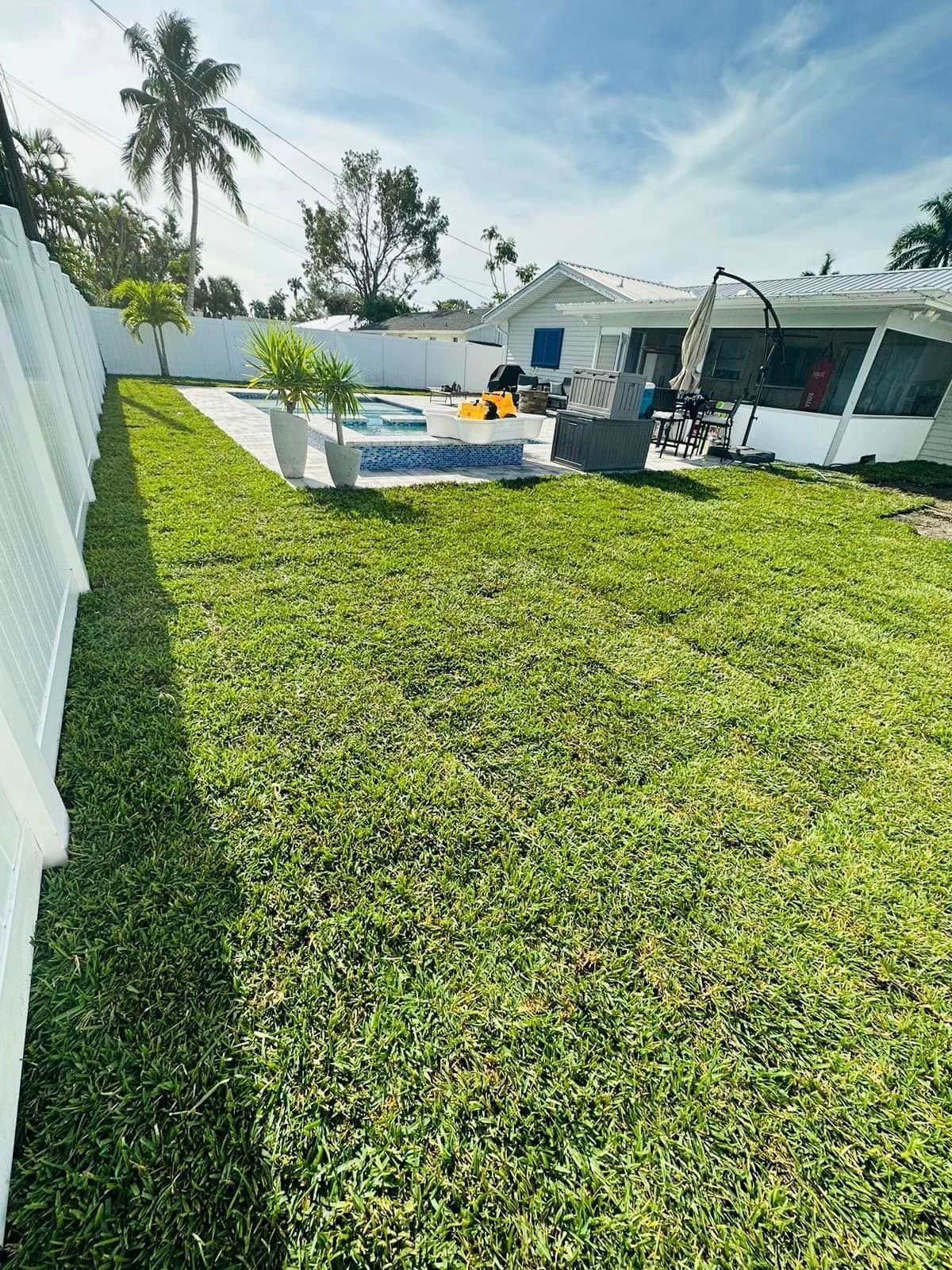 A backyard with a pool, green lawn, and white fence on a sunny day. A house is visible in the background.