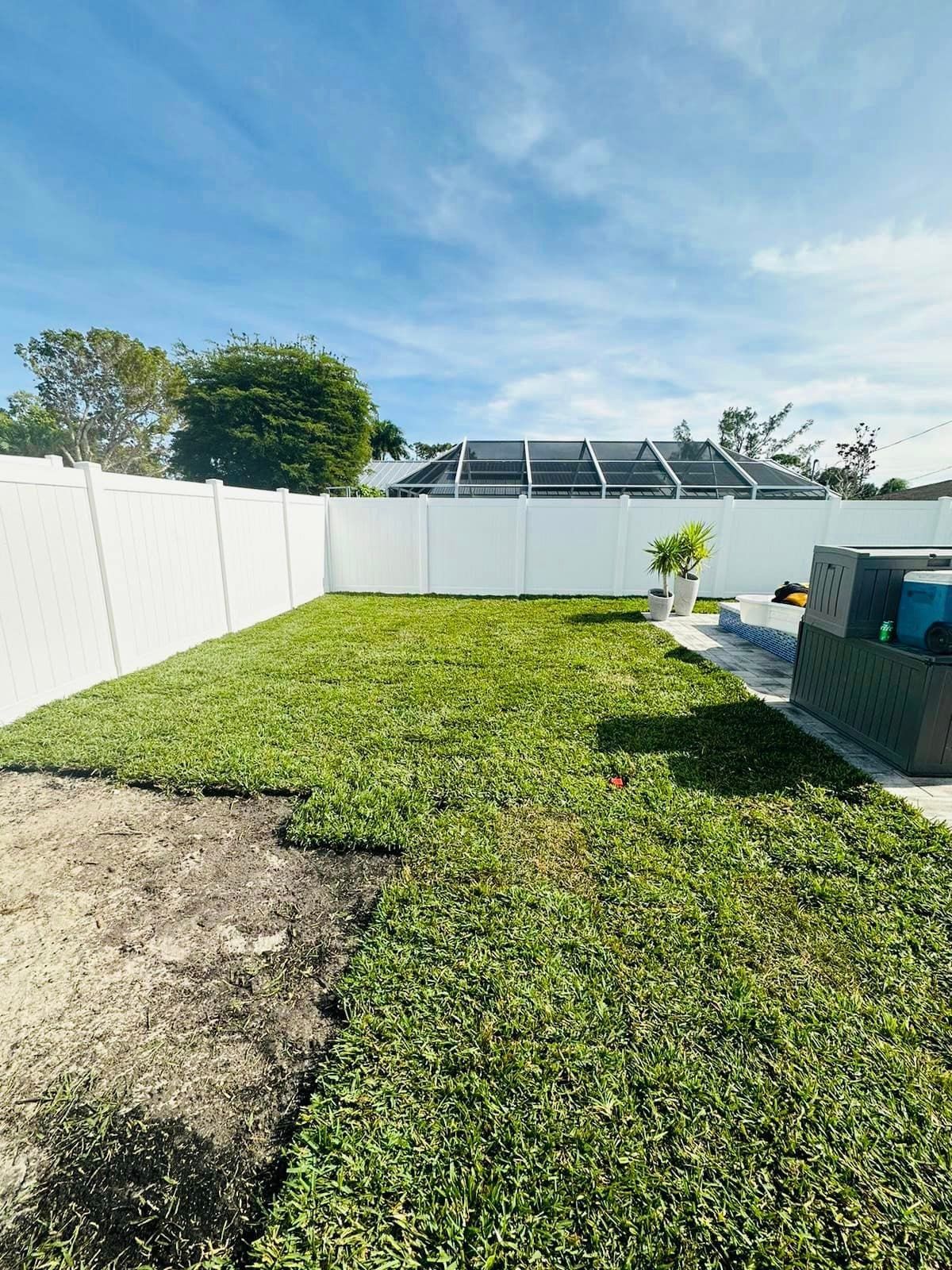 Green lawn and white fence in a backyard under a partly cloudy blue sky. Some new grass has been laid.