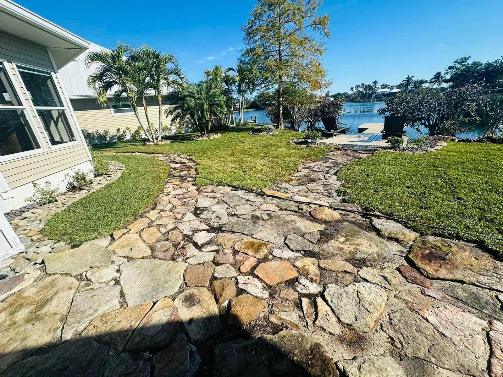 Stone pathway through a sunny backyard with grass and a view of the water, palms, and trees.