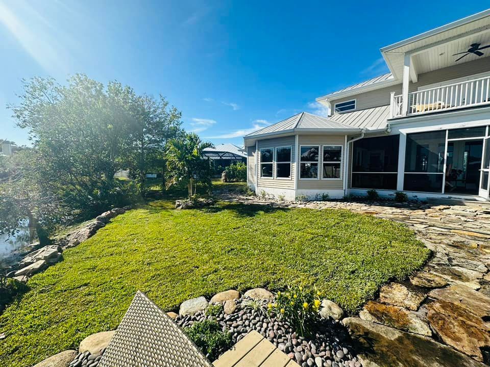 Lawn with a rock border by a canal, leading to a Florida-style house with screened porch on a sunny day.