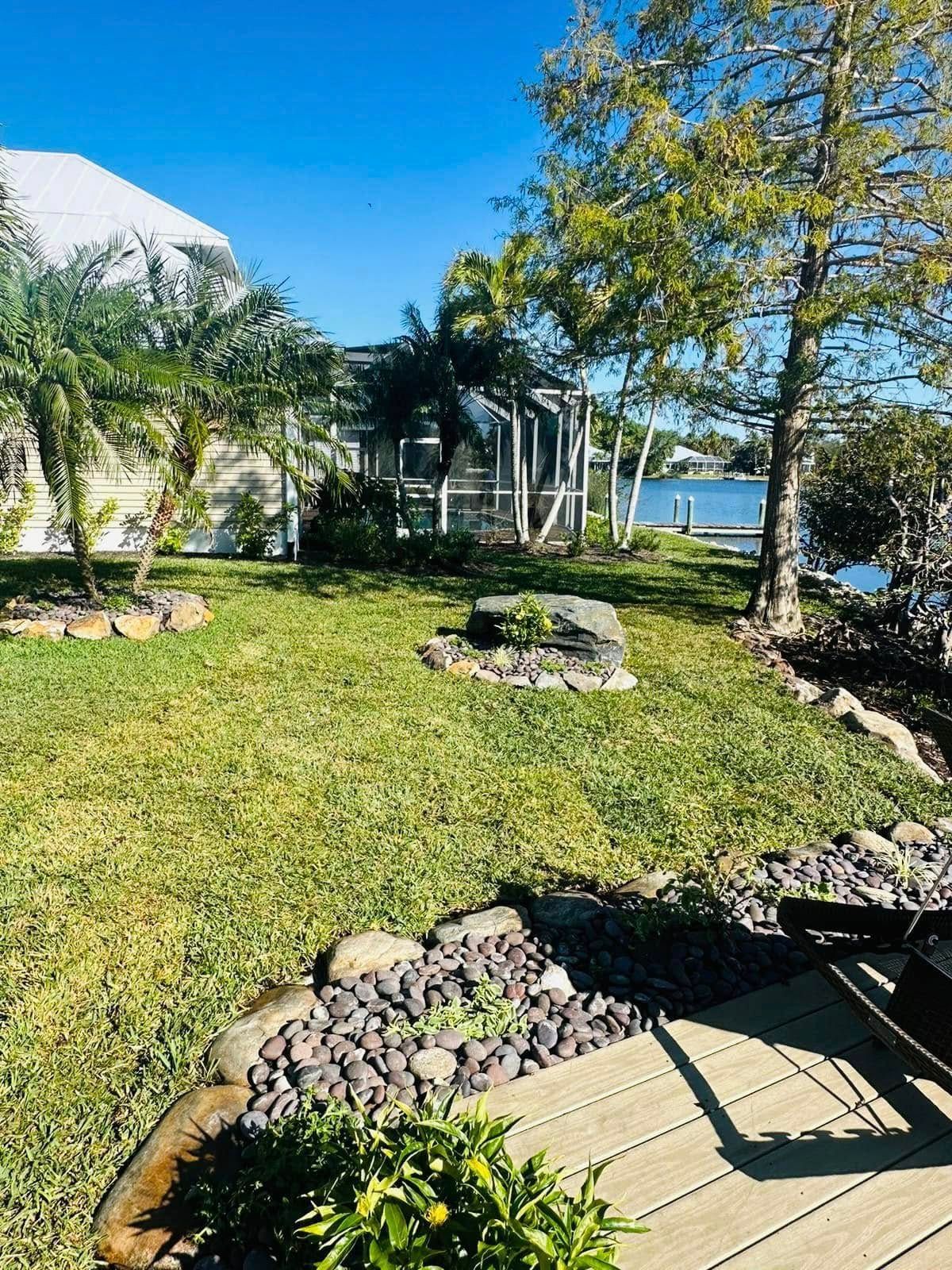 A sunny backyard with green grass, a rock garden, and a view of a lake.