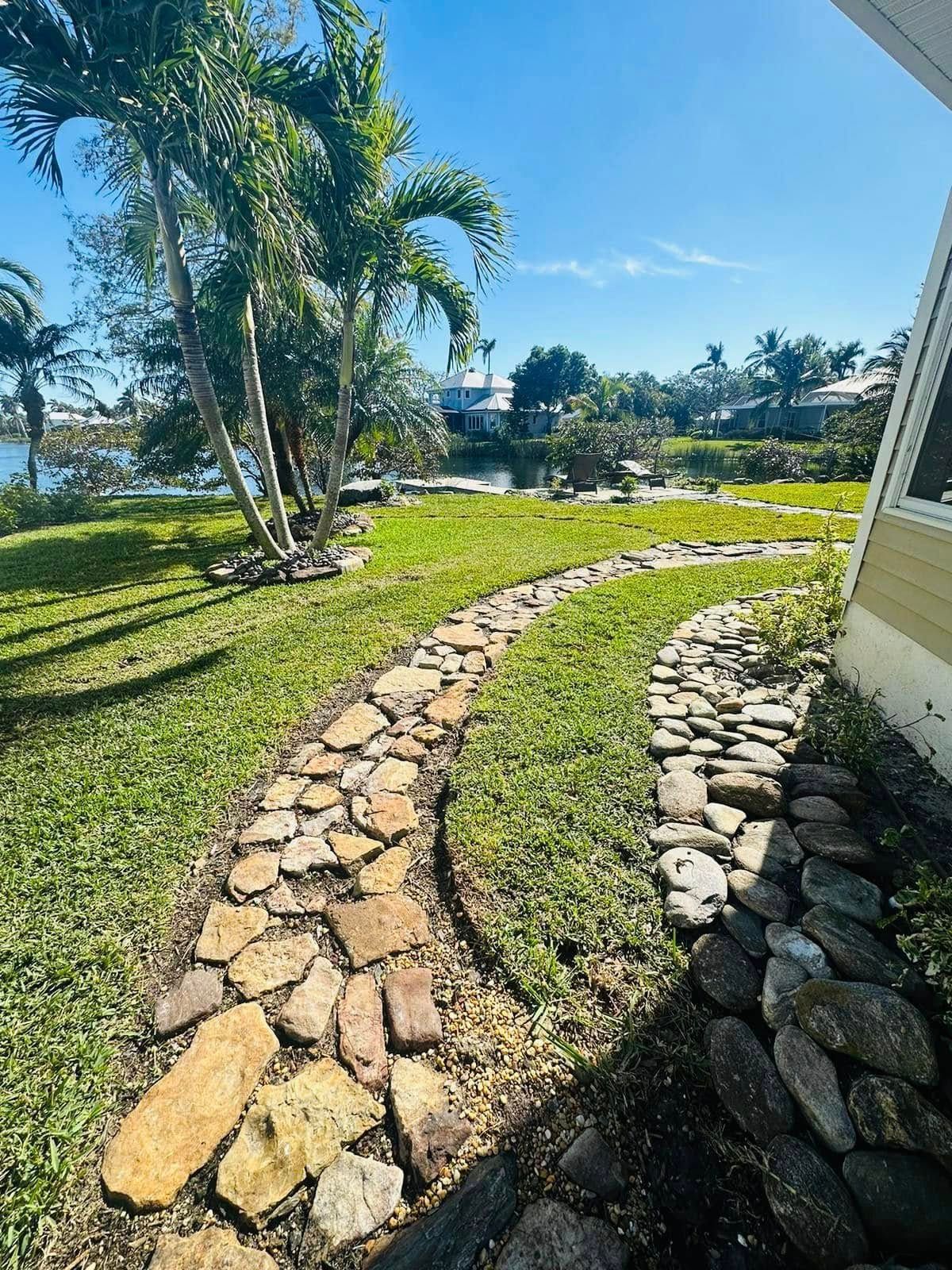 A stone path curves through a green yard towards a body of water, under a blue sky.