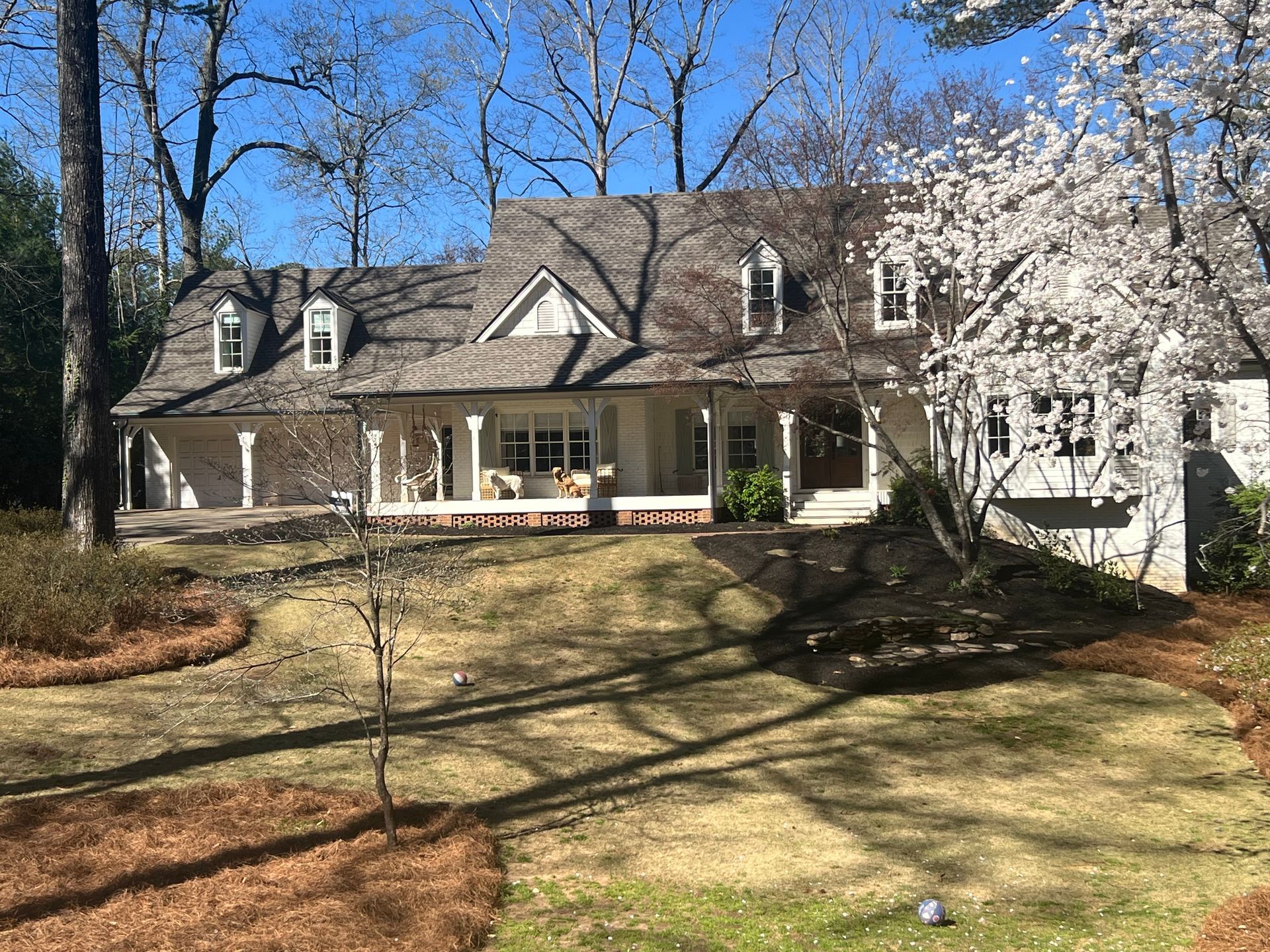 A large white house with a gray roof is surrounded by trees on a sunny day.