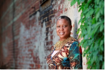 Woman smiling, leans against a brick and ivy wall, wearing a colorful patterned top with gold embellishments.