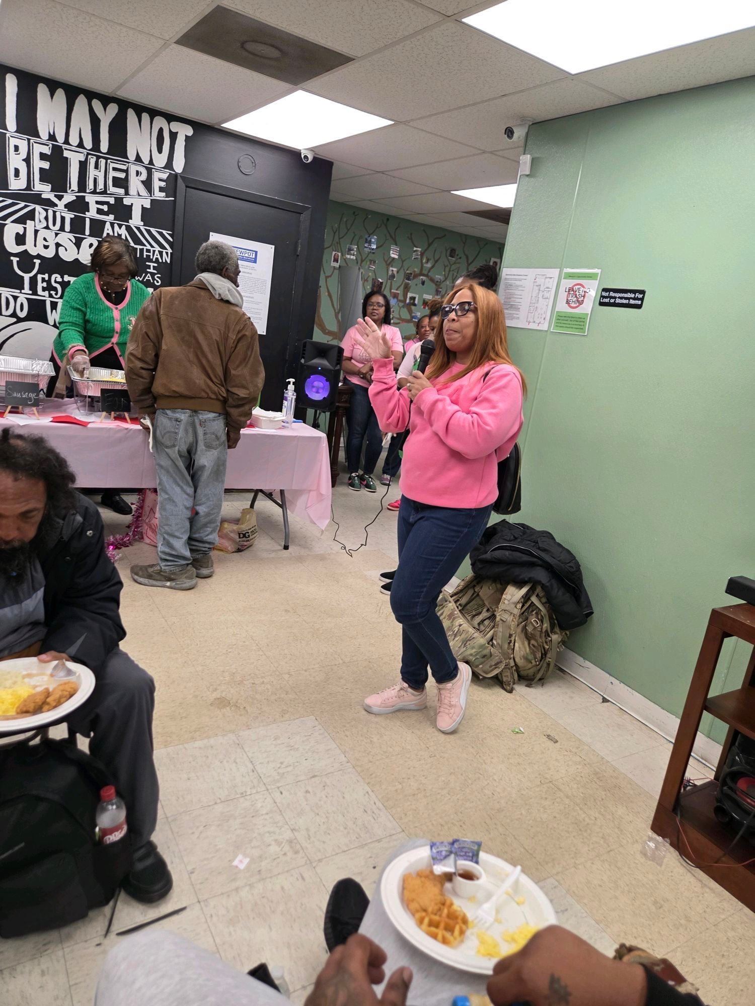 A woman in pink sings, others watch, and some eat at a table in a room.