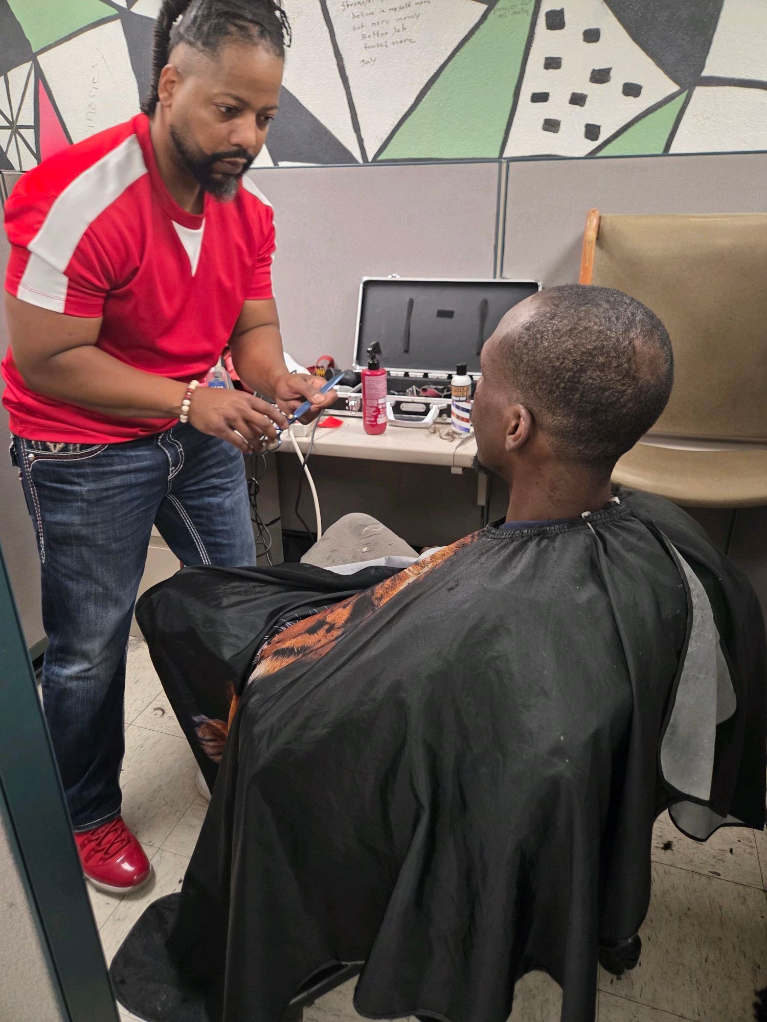 Barber giving a haircut to a client wearing a black cape, indoors. The barber is wearing a red shirt and jeans.
