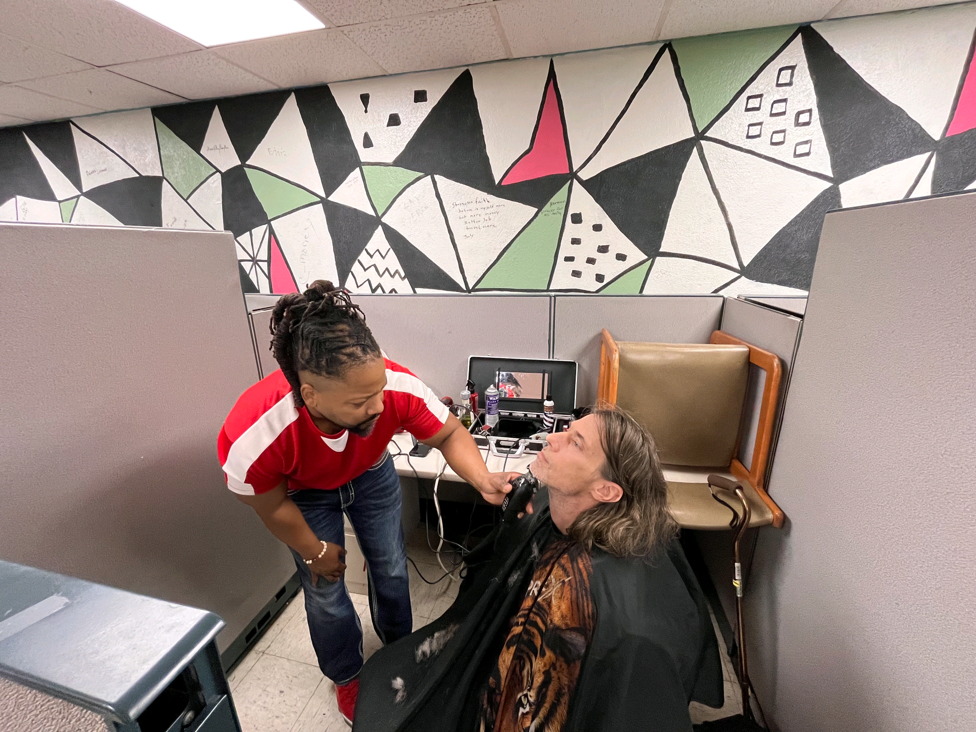 A person receives a haircut from another person in a room with geometric wall art.