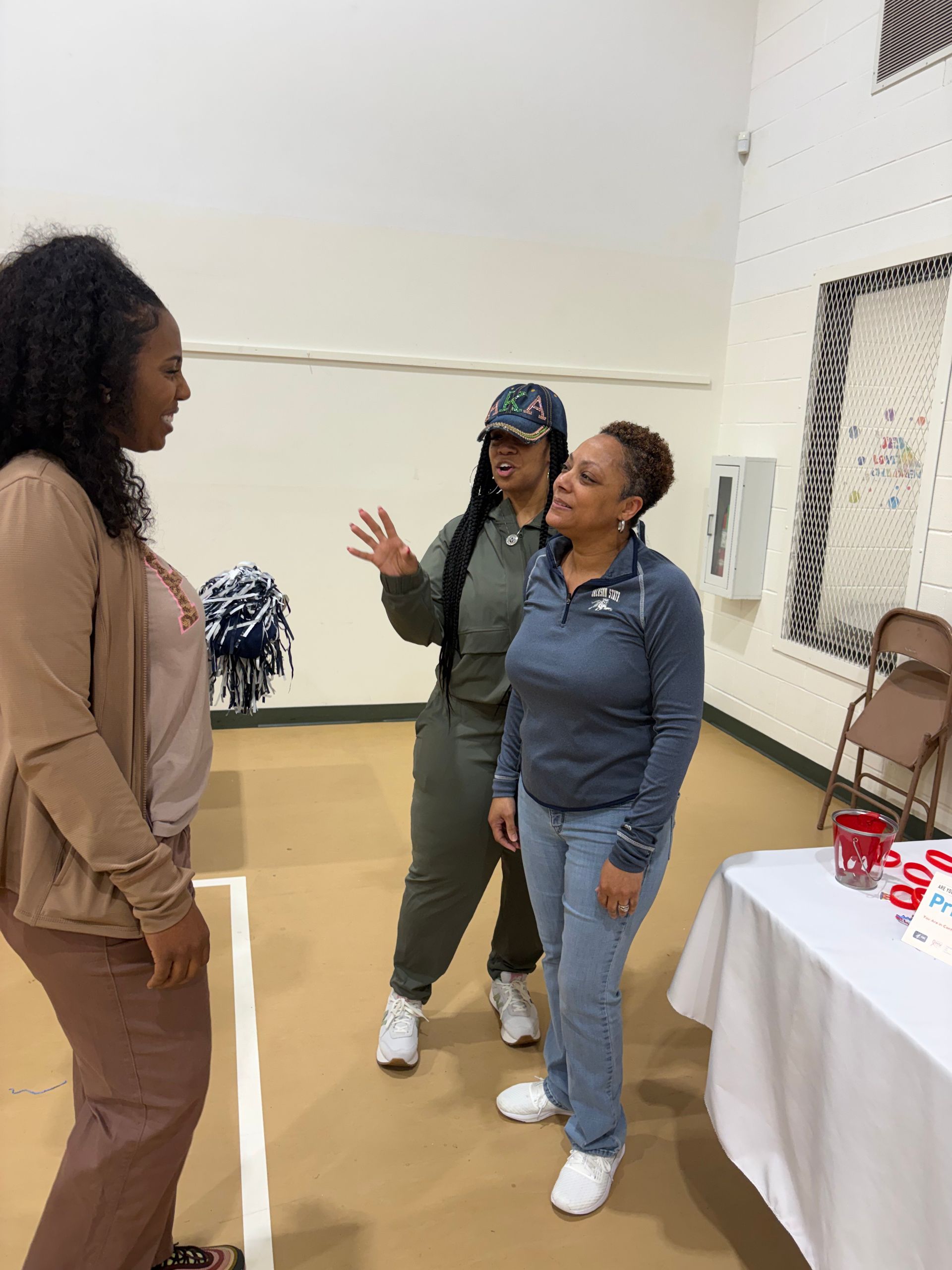 Three people talking near a table. Two women are gesturing, and one woman is listening, in a gymnasium setting.