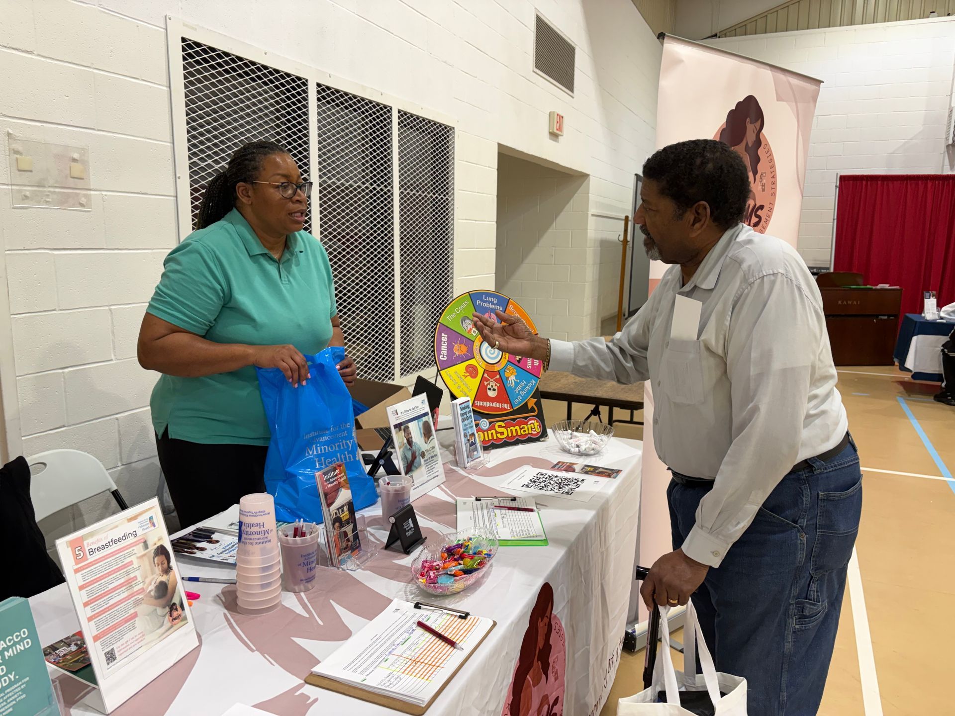 Woman at table talking with a man. Various items on table: flyers, gifts, and a prize wheel in a community center.