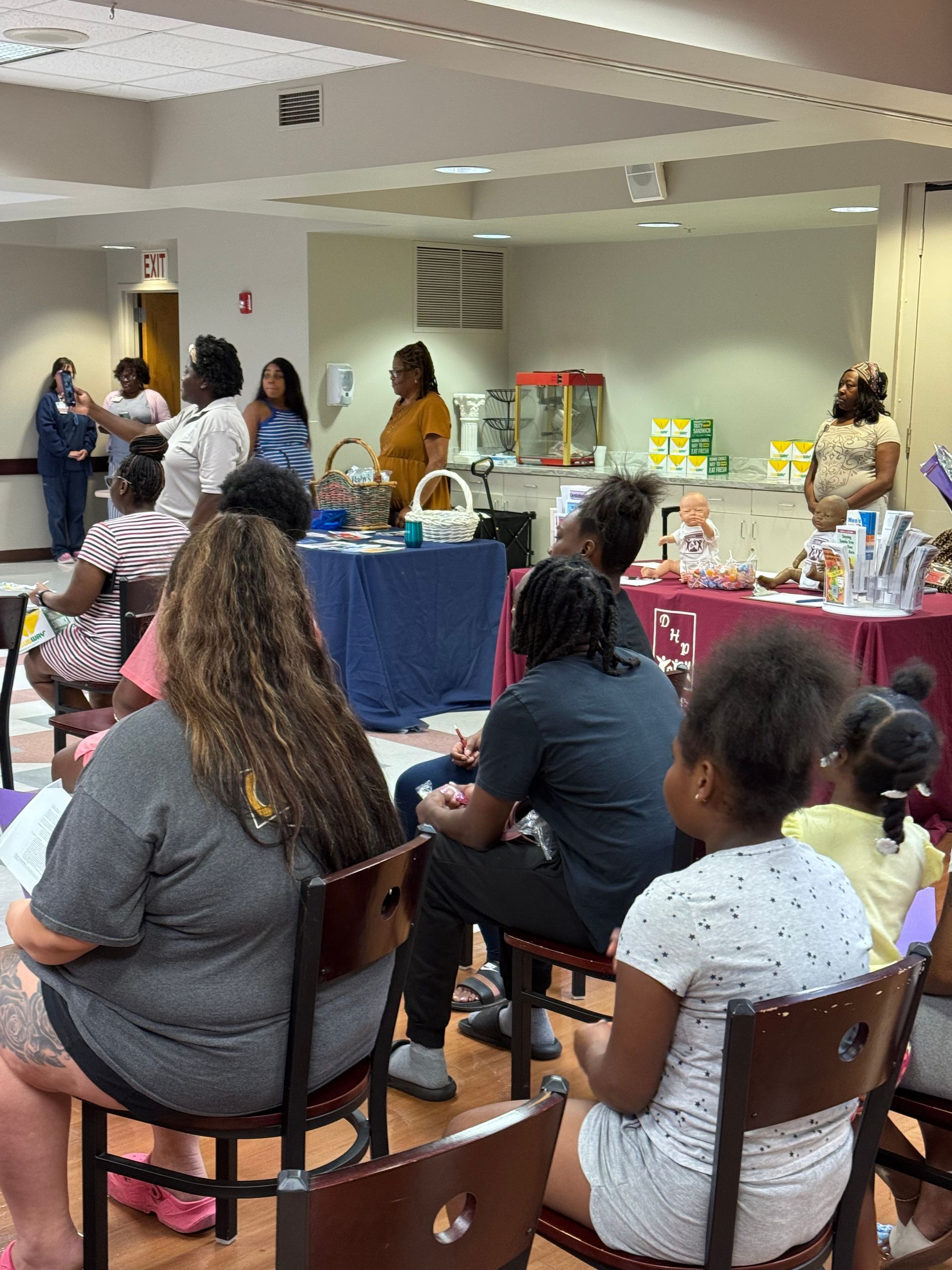 People gathered for an event. One woman speaks at a table with items. Others listen from chairs.