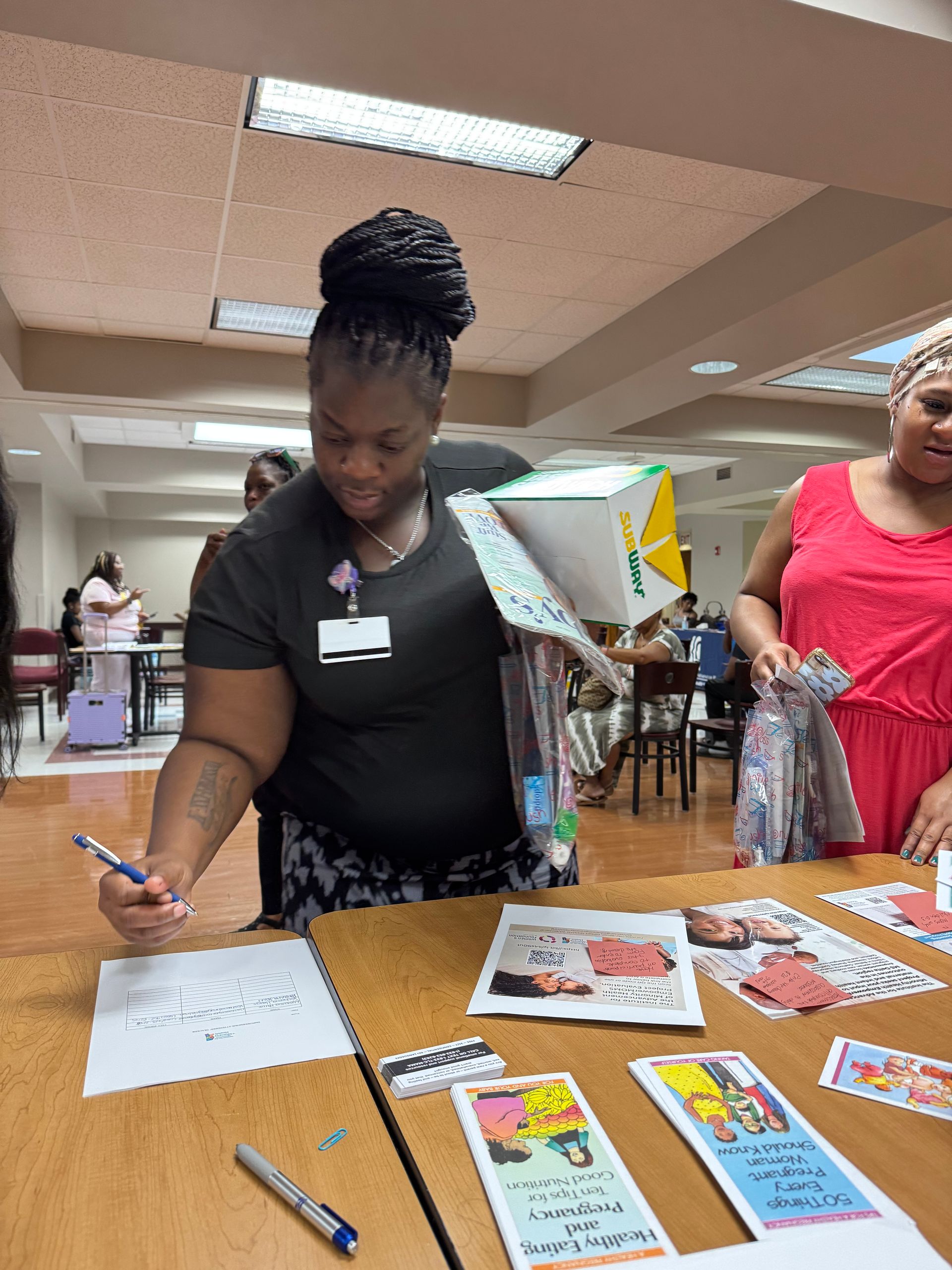 Woman writing at a table, holding a box with materials. Another woman stands nearby. Bright, indoor setting.