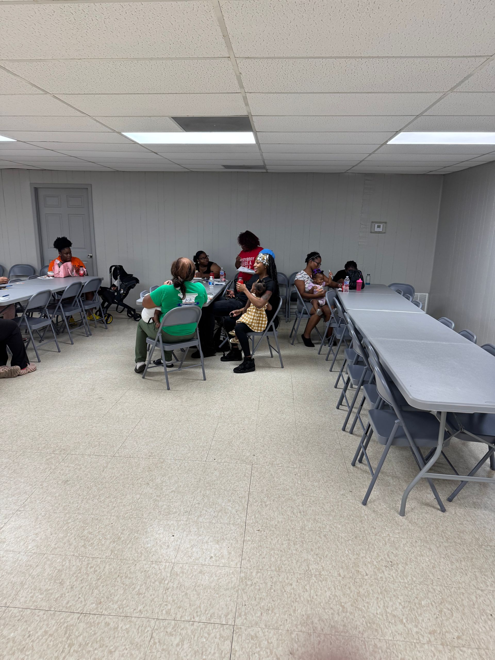 People seated at tables in a room. Some are talking, others look towards the camera. Gray tables and chairs. White walls and ceiling.