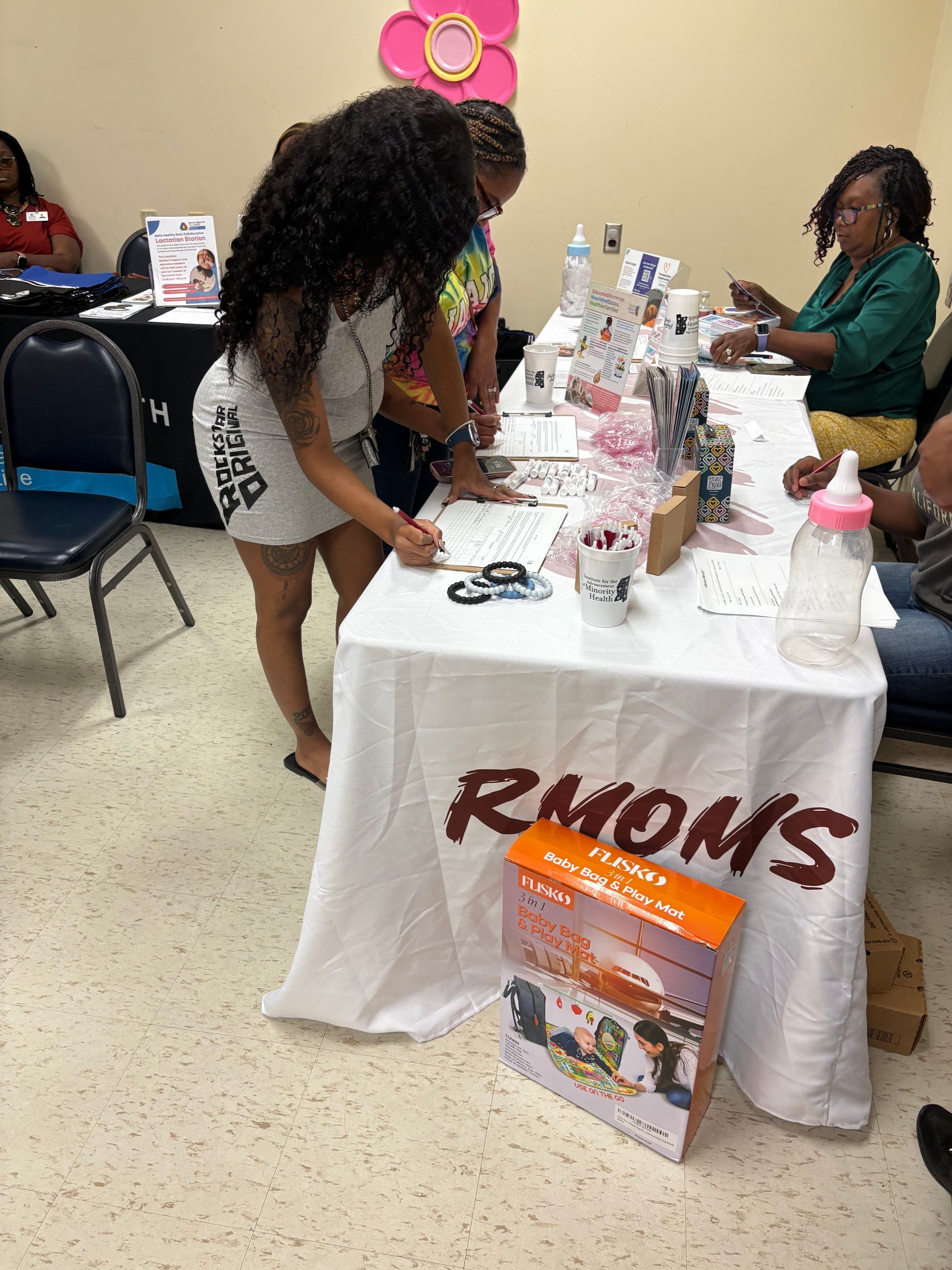 People at a table with "RMOMS" logo, signing papers, baby supplies on the table.