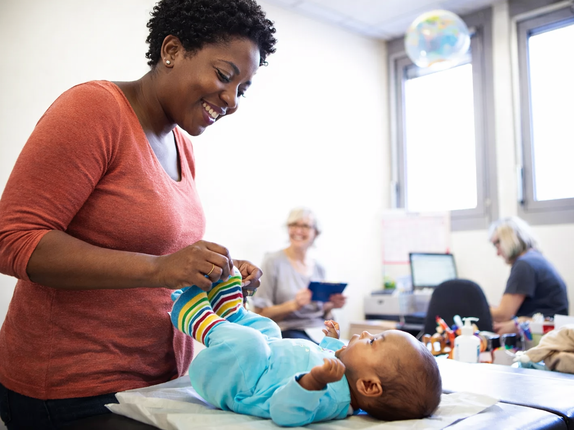 Doctor examining pregnant person's abdomen, smiling. Light-skinned person in pink shirt; setting appears to be a bedroom.