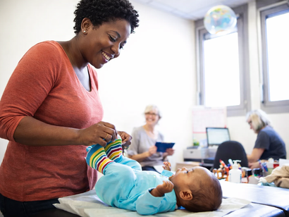 Doctor examining pregnant person's abdomen, smiling. Light-skinned person in pink shirt; setting appears to be a bedroom.