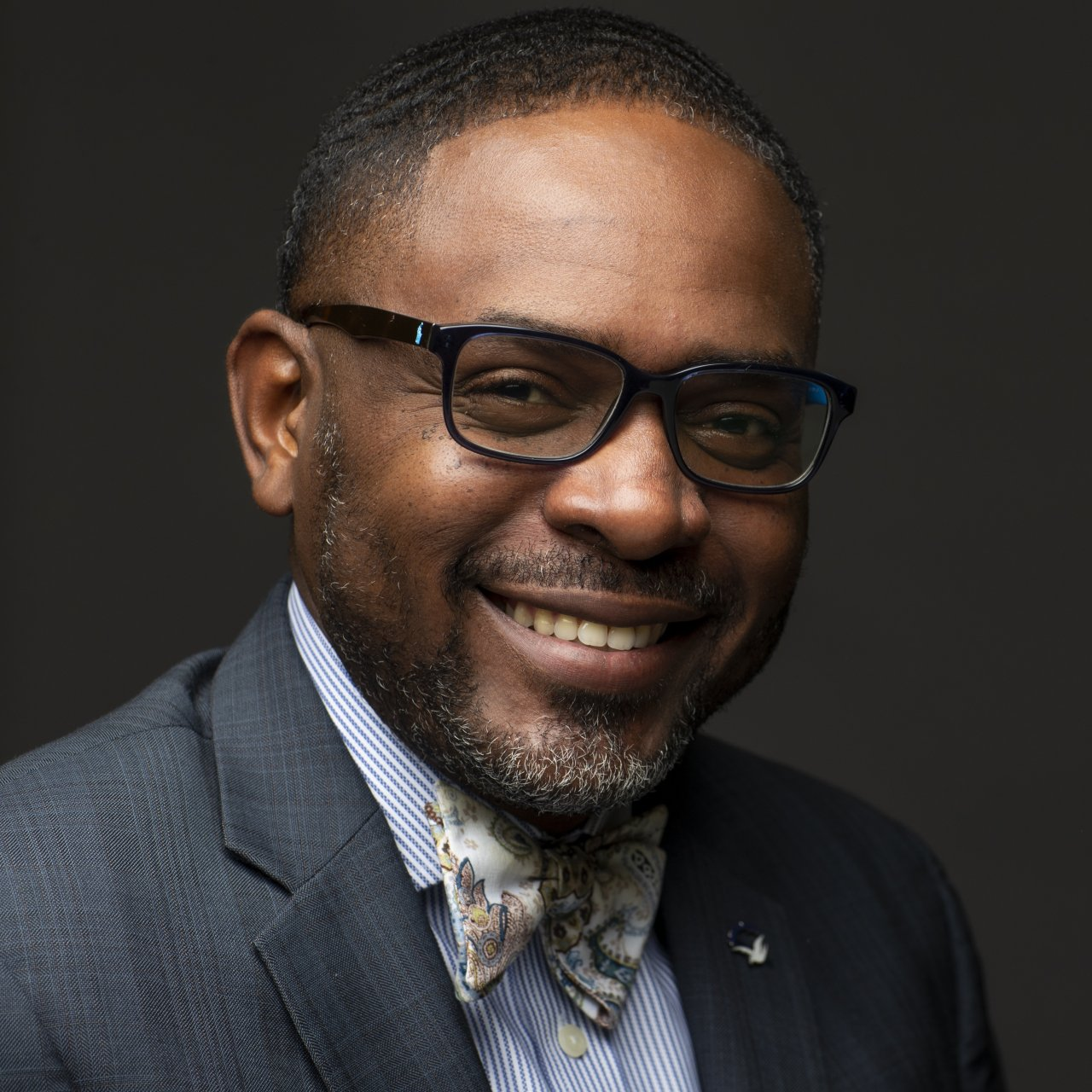 Man wearing glasses and patterned bow tie smiles, posed against a dark background.