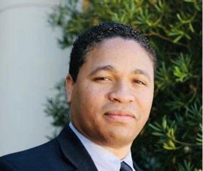 Man in dark suit, light skin, looking forward, soft smile, outdoor setting.