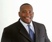 Man in a black suit with a blue patterned tie smiles in front of a white background.