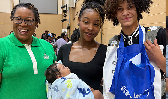 Three people posing indoors, one holding a newborn wrapped in a blue blanket and one holding a blue gift bag.