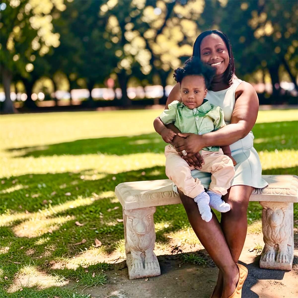 Woman holding a child on a stone bench in a sunlit park, both smiling at the camera.