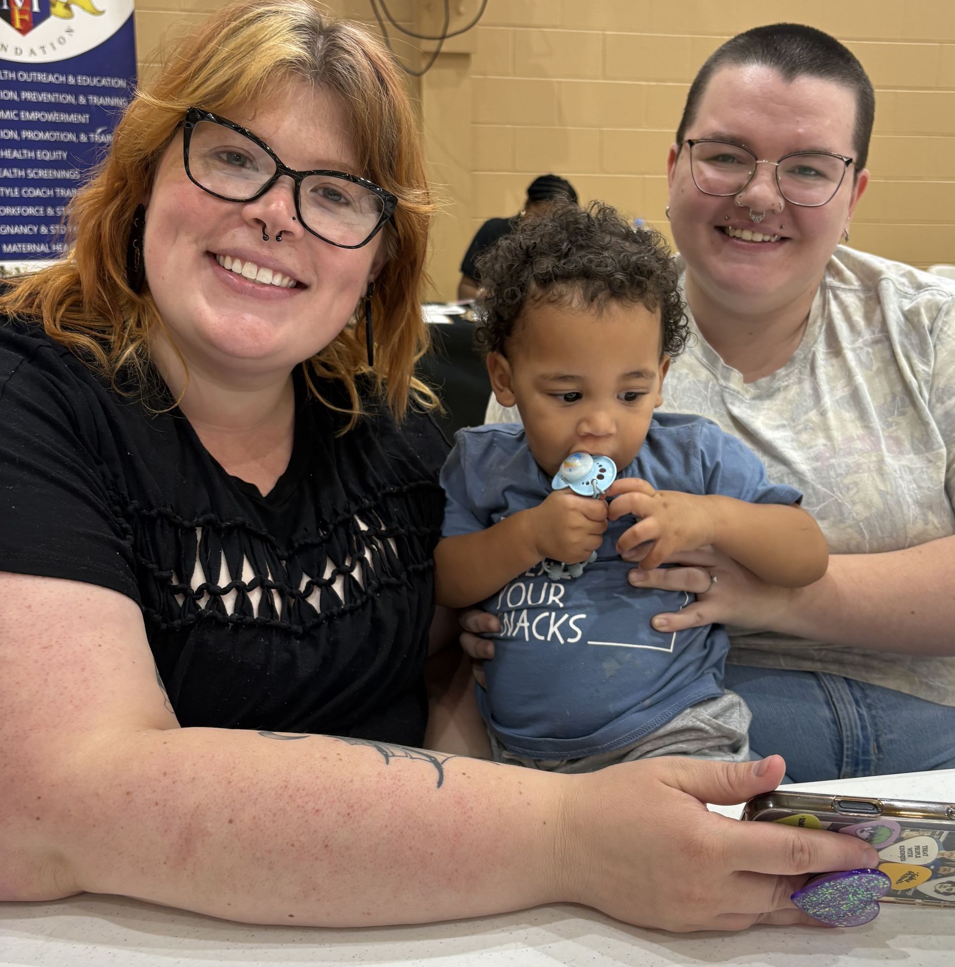 Three people smiling at a table, with a child holding a snack in the center.