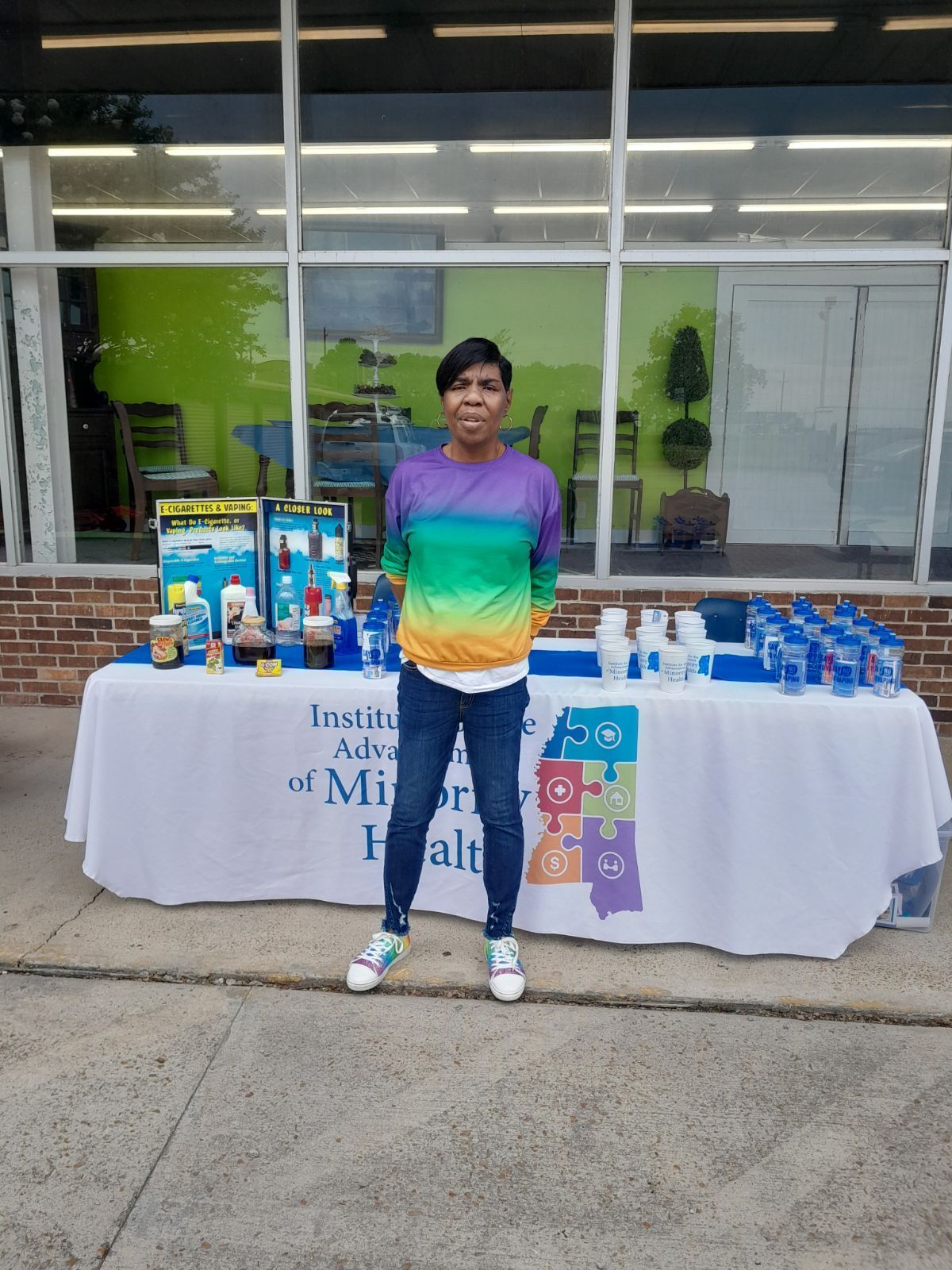 Woman stands at a table with drinks and water bottles, in front of a building with a white and green wall.