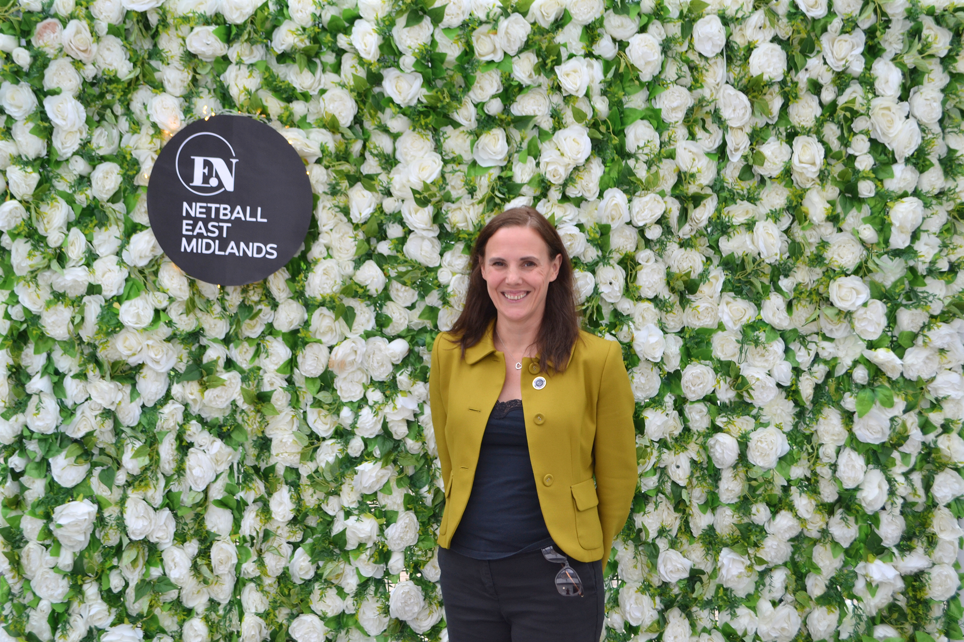 A woman in a yellow jacket is standing in front of a wall of white flowers.