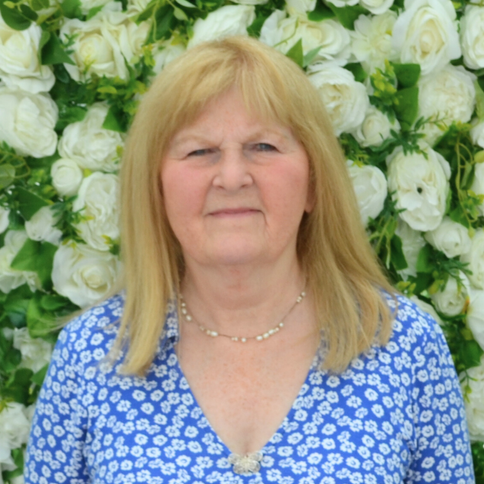 A woman in a blue shirt is standing in front of a wall of white flowers.