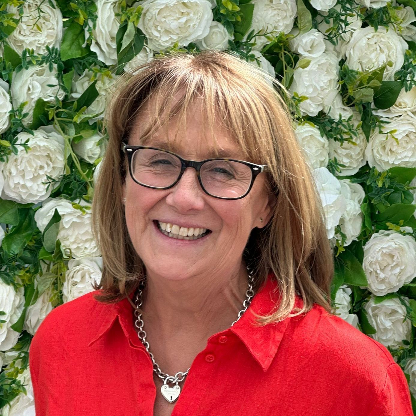 A woman is smiling in front of a wall of white flowers